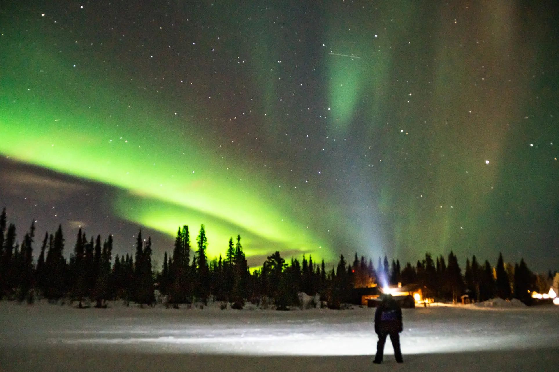 person watching aurora borealis over snowy forest and cabins