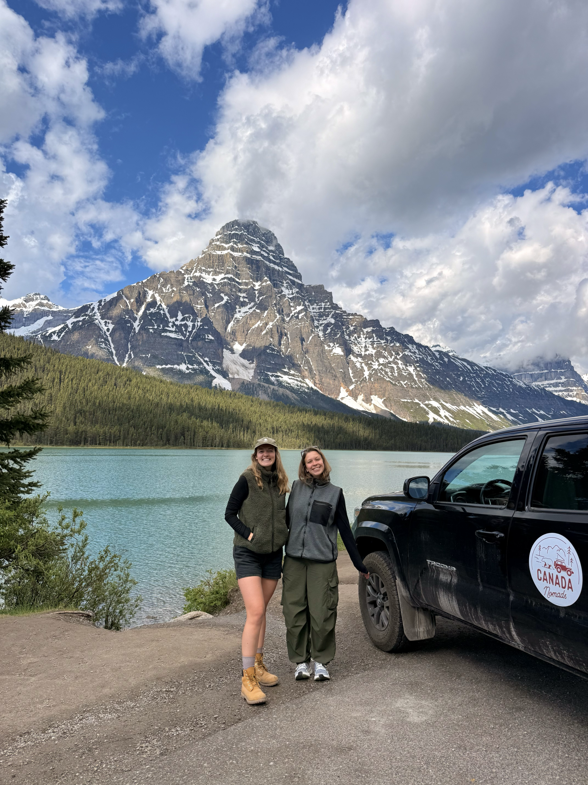 two women posing with a truck by a turquoise lake and mountains